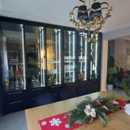 Nowoczesna, czarna witryna z podświetleniem LED, wypełniona szkłem, alkoholem i dekoracyjnymi figurkami, widoczna zza stołu z świąteczną dekoracją w salonie.