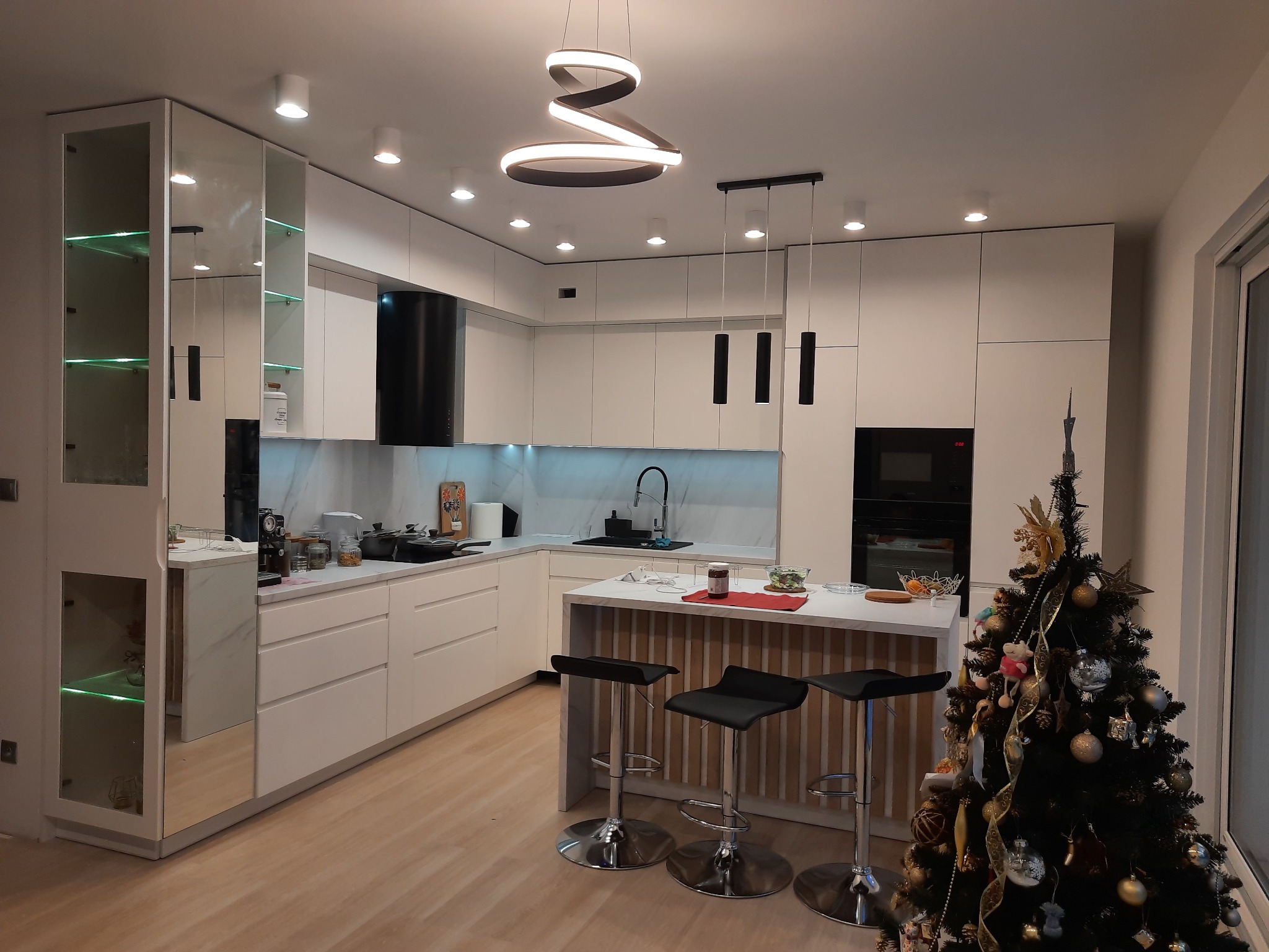 Nowoczesna, jasna kuchnia z wyspą i hokerami, oświetlona spiralną lampą. Świąteczny akcent w postaci choinki przy oknie. Fronty lakierowane, system BLUM.