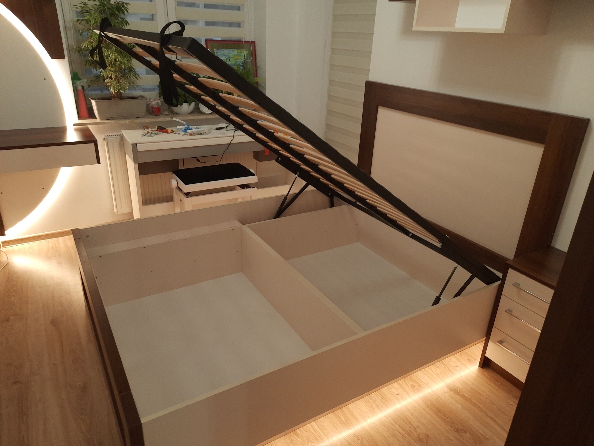 Otwarta rama łóżka z pojemnikiem, widoczne wnętrze z podziałem na dwie części, podświetlenie LED na podłodze, zagłówek z drewnianą ramą, szafka nocna obok.