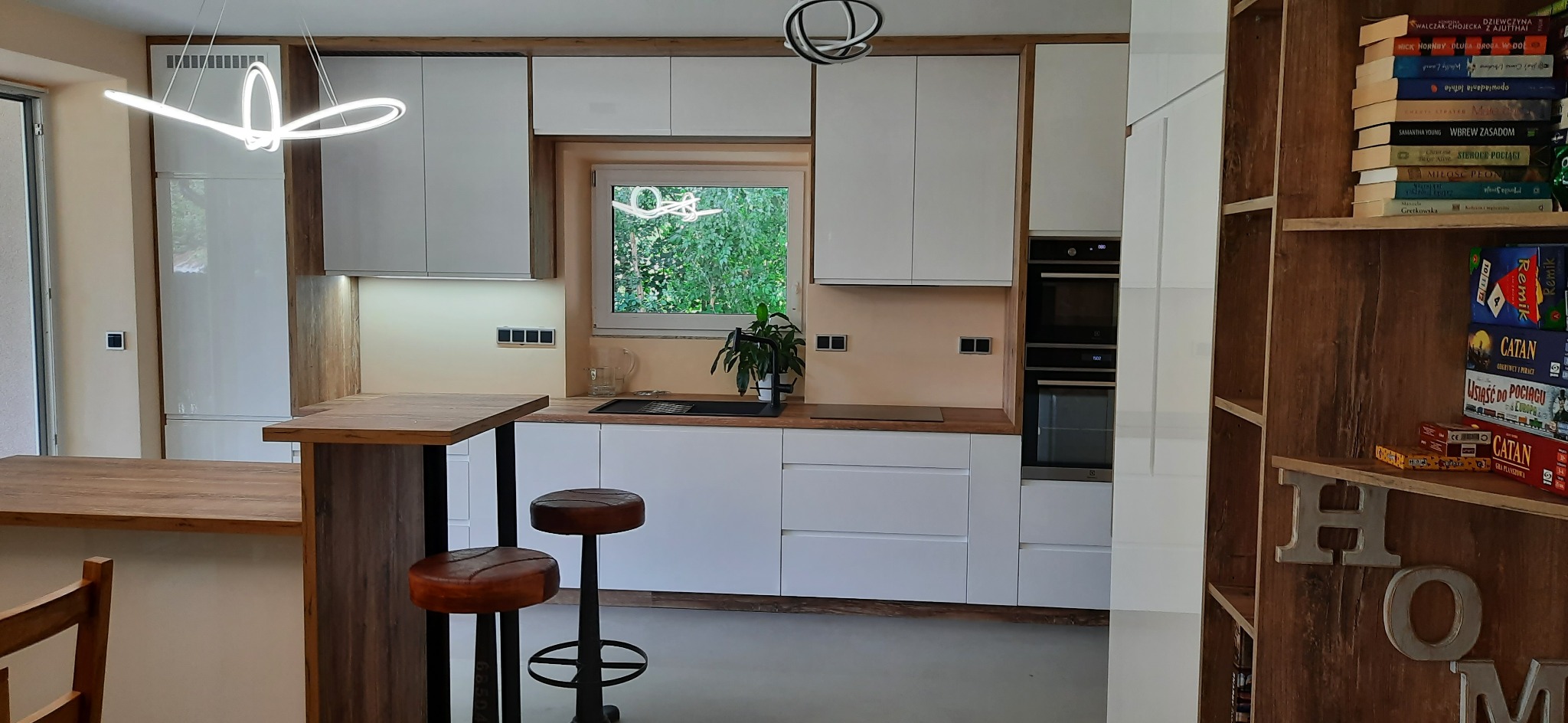 Jasna kuchnia z białymi szafkami i drewnianymi akcentami, wyspą z hokerami i oknem z widokiem na zieleń. Nowoczesne oświetlenie i regał z książkami.