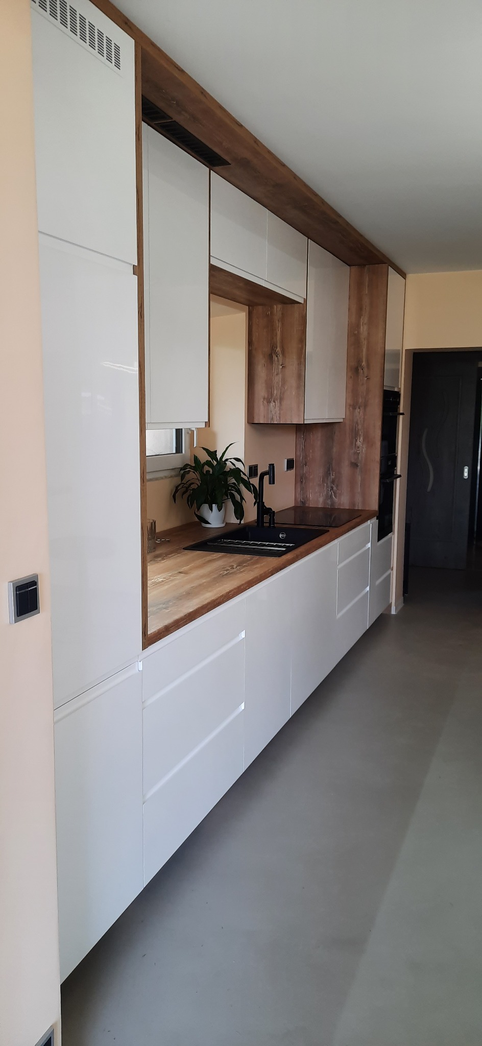 Nowoczesna kuchnia z białymi, błyszczącymi frontami szafek, drewnianym blatem i wykończeniem w kolorze drewna. Widoczny zlew, bateria i roślina w doniczce.