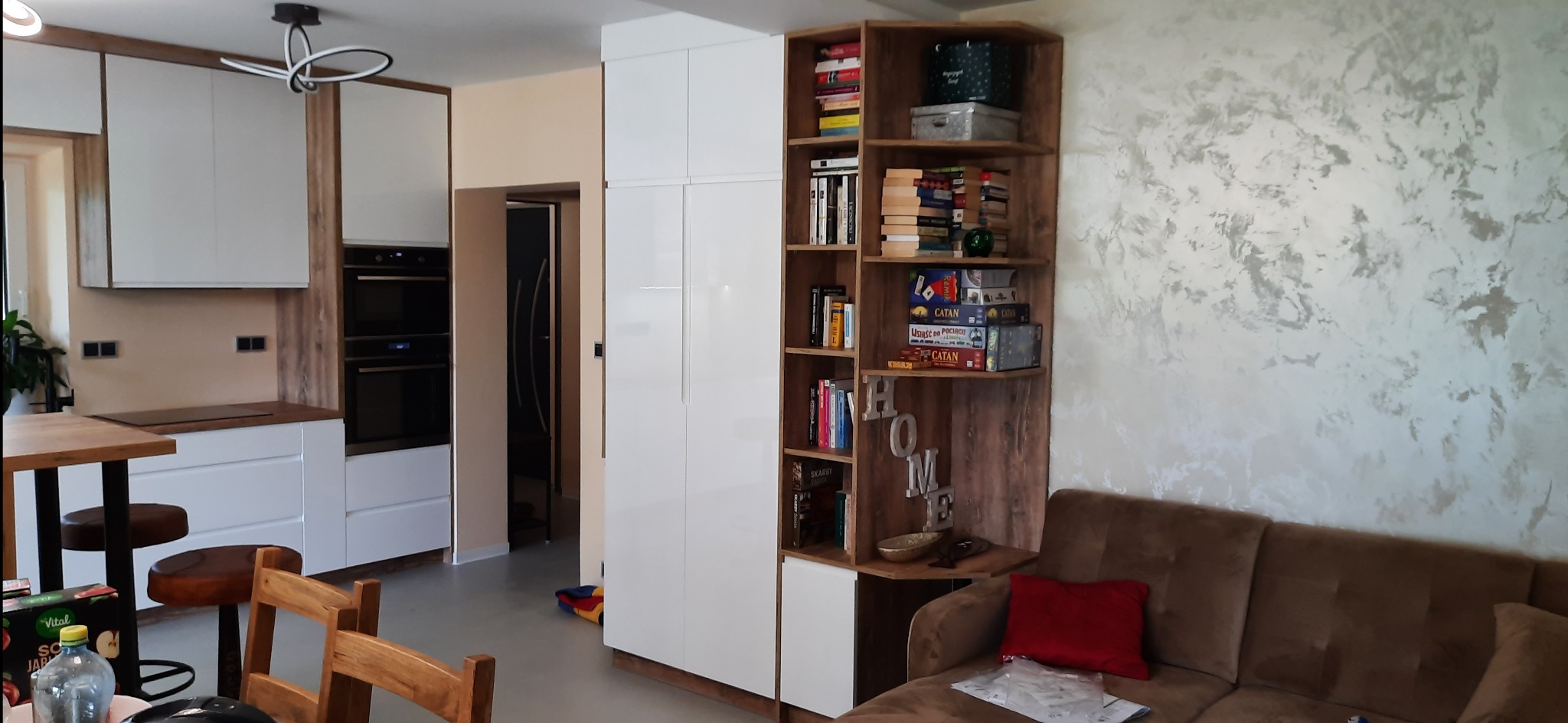 Nowoczesna kuchnia z białymi szafkami i drewnianymi akcentami, połączona z strefą relaksu z regałem narożnym i sofą. Widoczne gry planszowe na półkach.