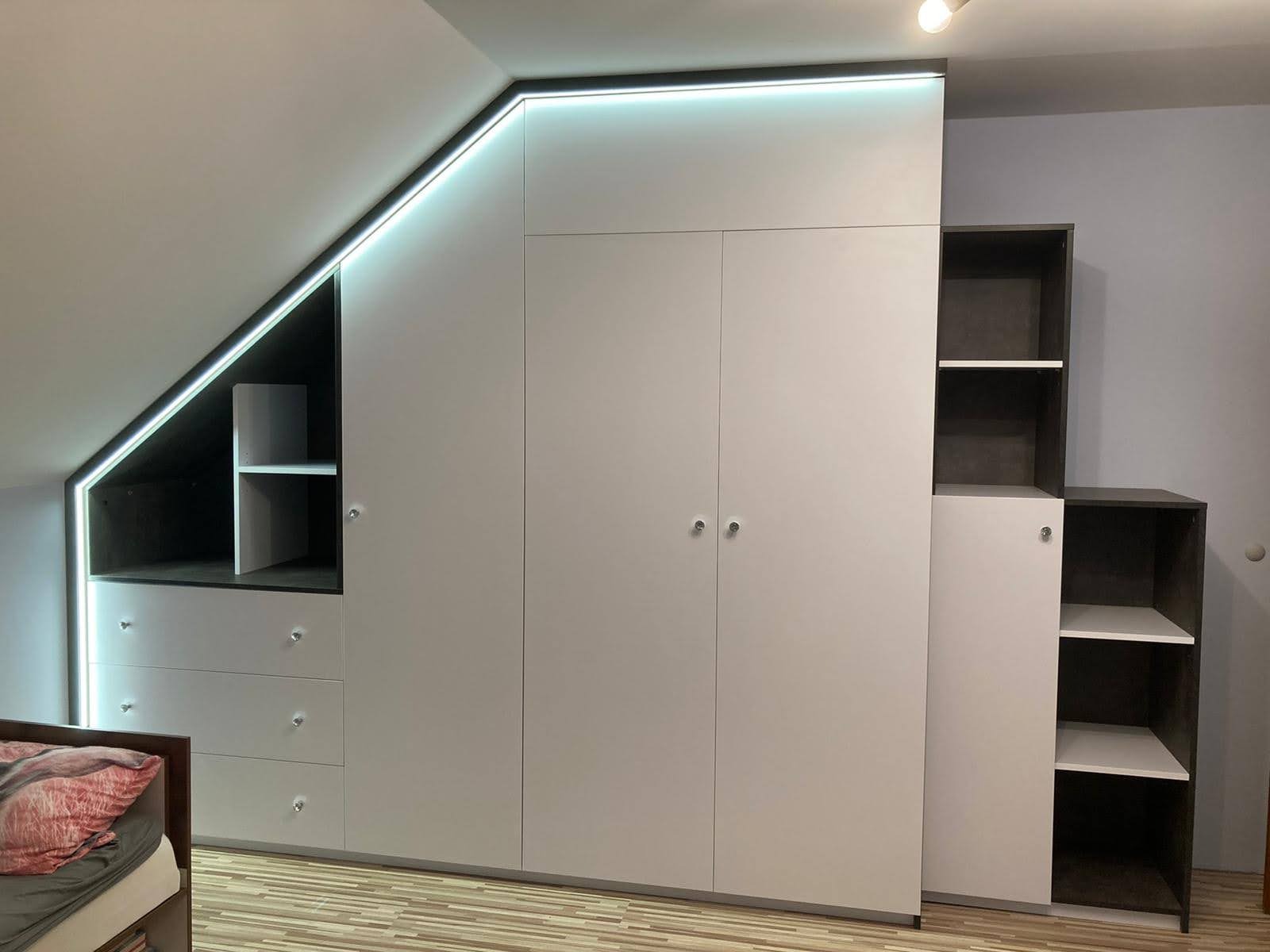 Biała szafa wnękowa z oświetleniem LED, idealnie dopasowana do skosu poddasza. Widoczne szuflady i półki, z minimalistycznymi, okrągłymi uchwytami.
