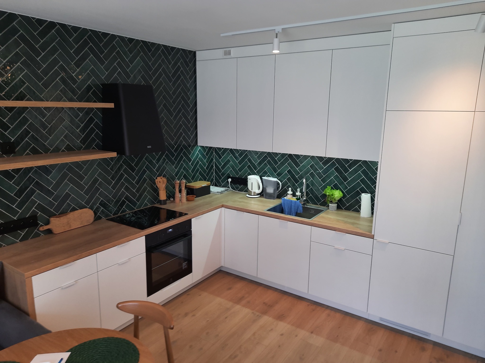 Nowoczesna kuchnia w kształcie litery L z białymi frontami, drewnianym blatem i zielonymi płytkami ułożonymi w jodełkę. Widoczny okap i półki z drewna.