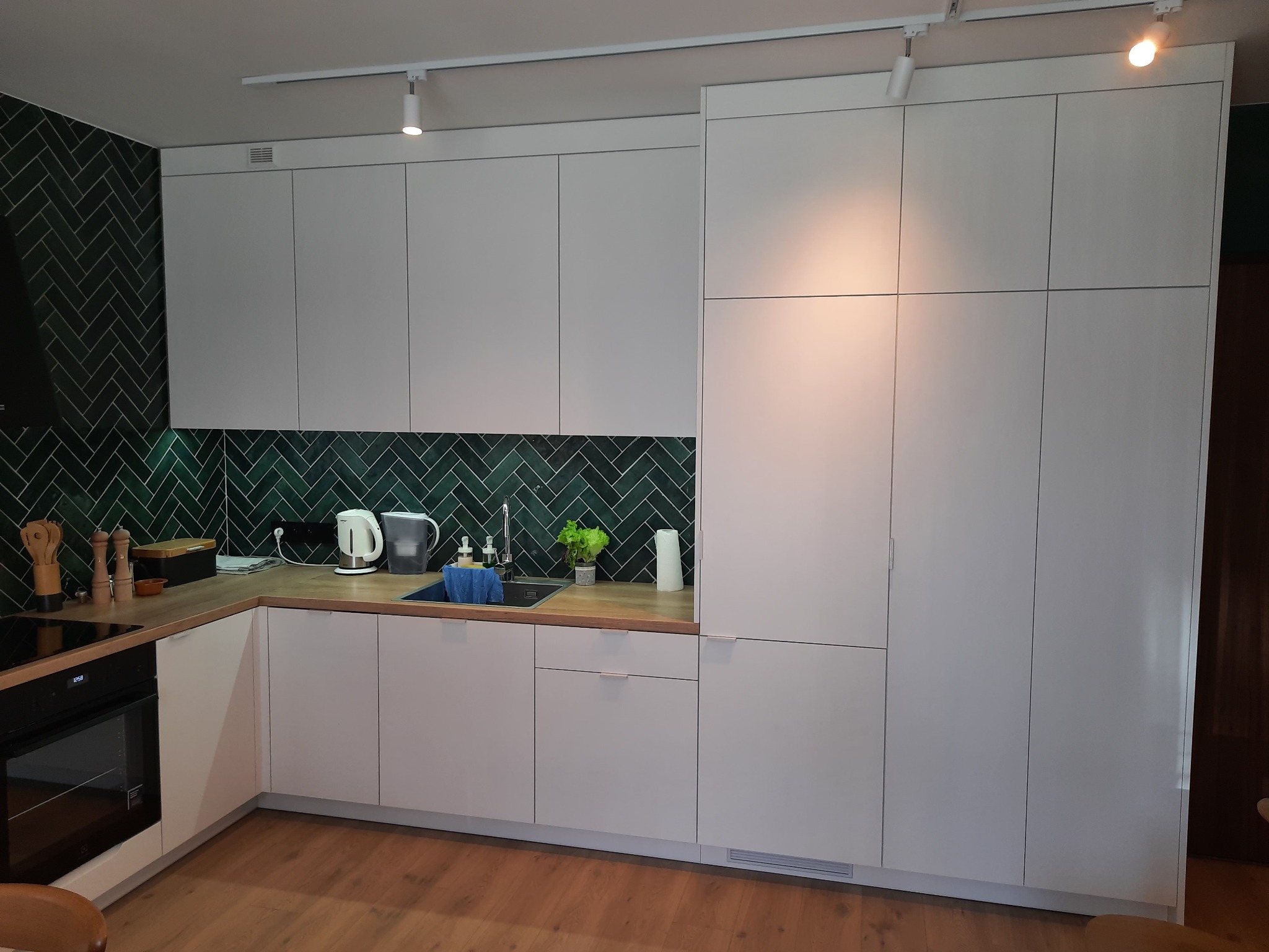 Nowoczesna kuchnia z białymi frontami i blatem drewnopodobnym, zielone płytki ułożone w jodełkę, oświetlenie punktowe. Minimalistyczny design, funkcjonalna zabudowa.