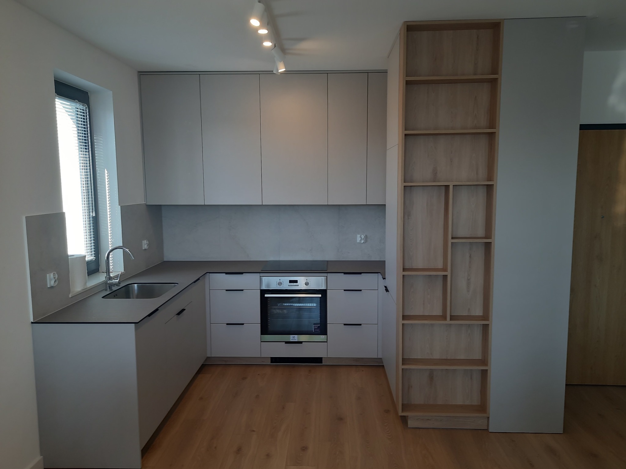 Jasna kuchnia z szarymi frontami i czarnym blatem, oświetlenie punktowe, drewniana podłoga i regał z jasnego drewna. Minimalistyczny design.