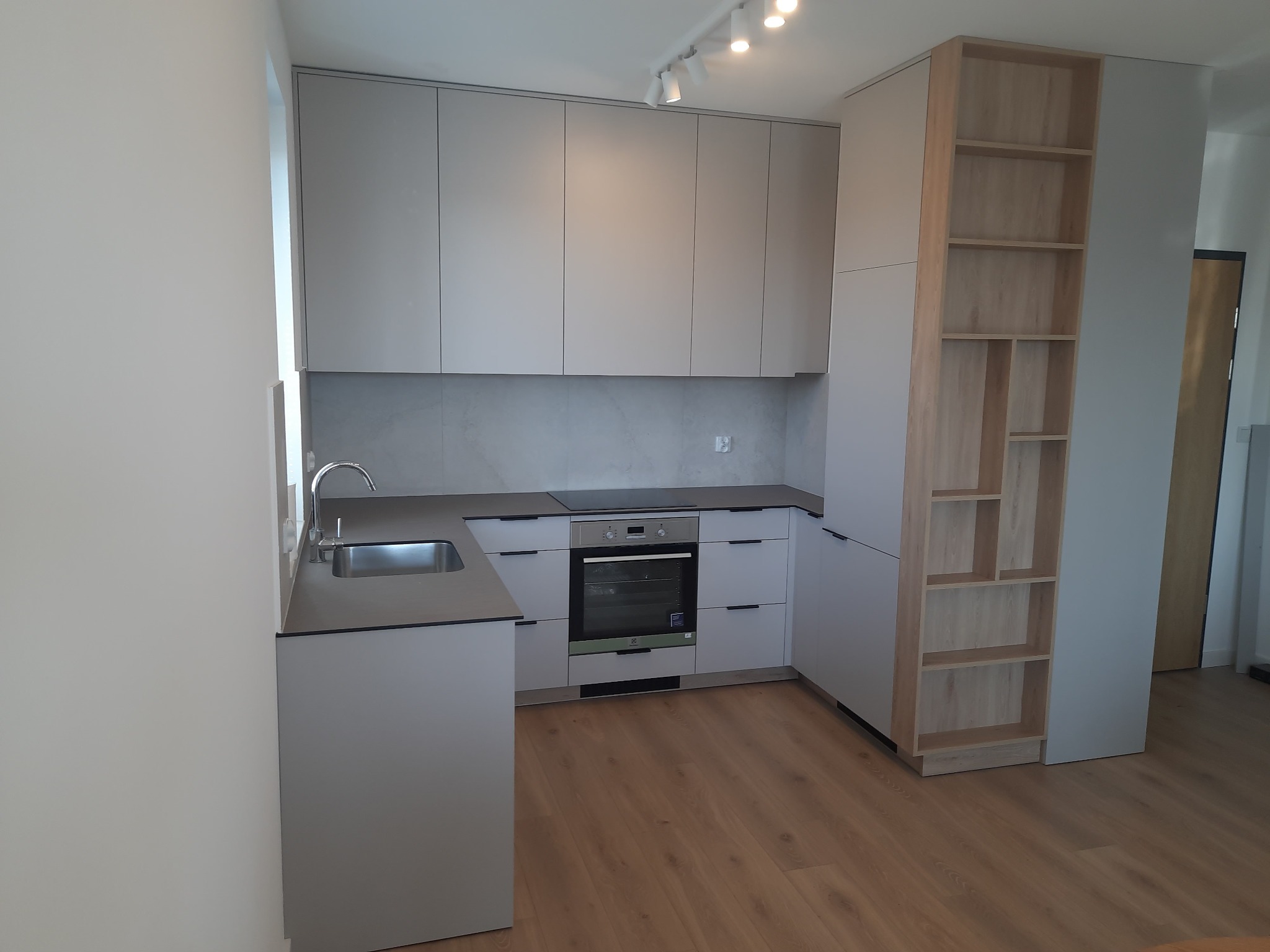 Jasnoszara kuchnia na wymiar z drewnianym regałem w zabudowie, zlewem i płytą indukcyjną. Minimalistyczny styl, oświetlenie punktowe, drewniana podłoga.