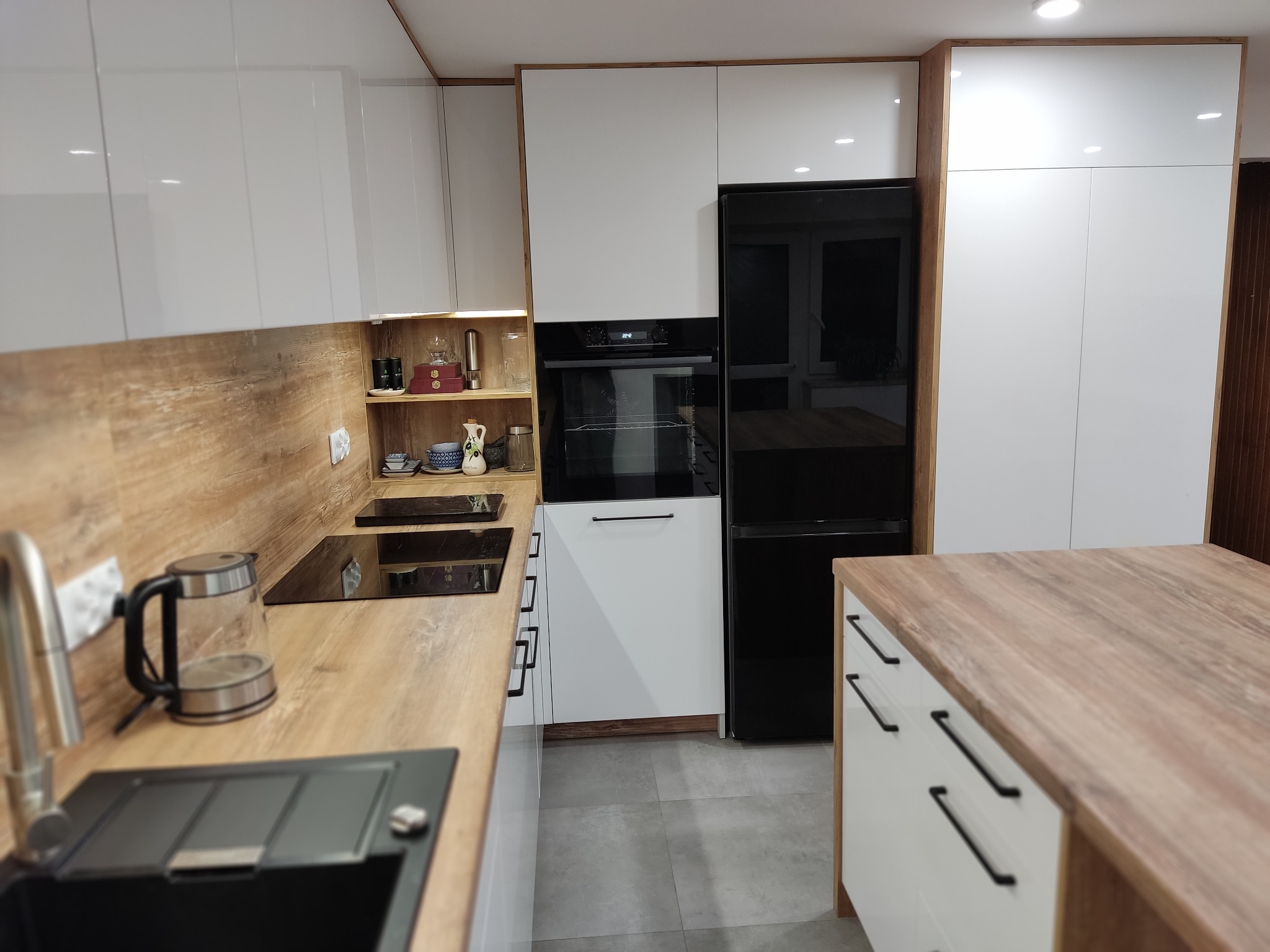 Nowoczesna kuchnia z białymi, lakierowanymi szafkami, drewnianym blatem i czarnymi akcentami. Widoczny zlew, płyta indukcyjna i wyspa kuchenna.