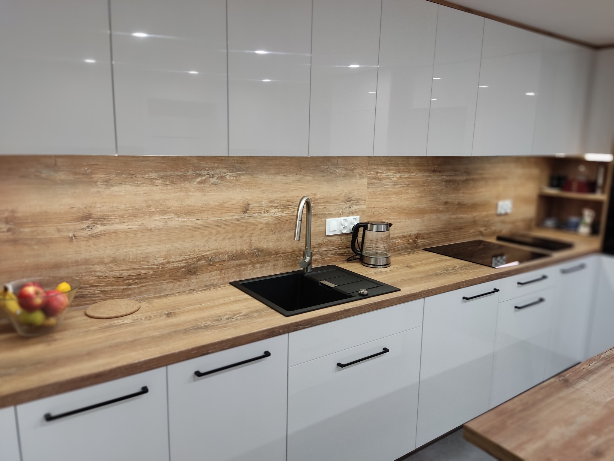 Nowoczesna kuchnia: białe szafki z czarnymi uchwytami, drewniany blat i panel ścienny, czarny zlew i minimalistyczna armatura. Owoce w szklanej misie.