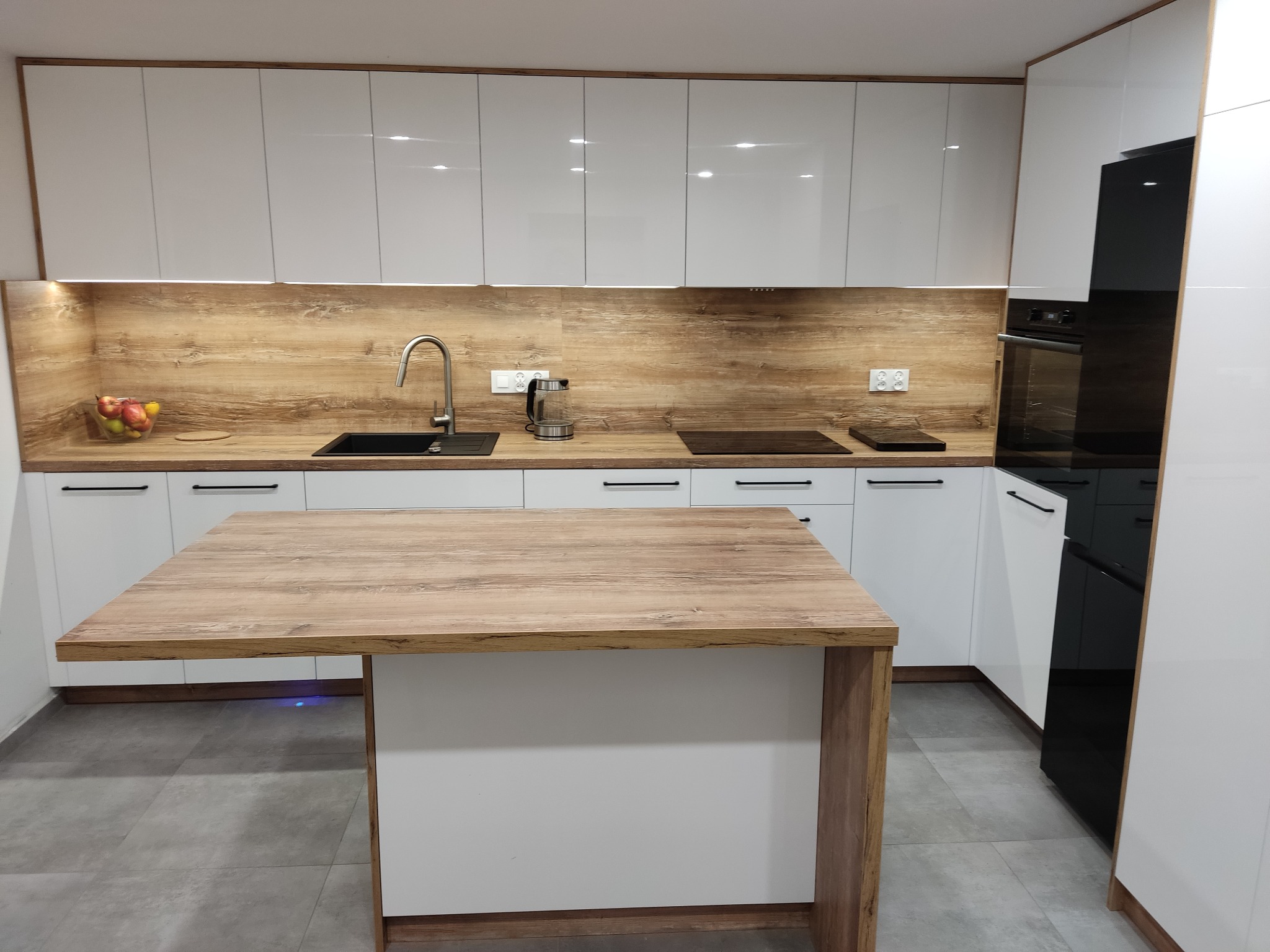 Nowoczesna kuchnia z białymi szafkami, drewnianym blatem i wyspą. Uwagę zwraca podświetlenie LED pod wyspą i minimalistyczne czarne uchwyty szafek.