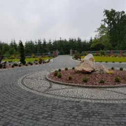 Szeroki, okrągły podjazd wyłożony szarą kostką brukową z dekoracyjnym wzorem z jasnej kostki, centralnie umieszczony okrągły klomb z brązowym żwirem i dużymi kamieniami, otoczony niskimi krzewami...