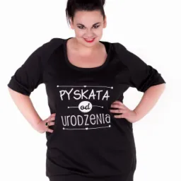 Kobieta pozuje w czarnej tunice z nadrukiem 'Pyskata od urodzenia', ręce oparte na biodrach, pierścionek na palcu.