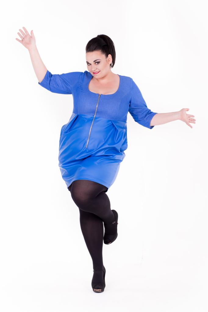 Kobieta w niebieskiej sukience z zamkiem i czarnych rajstopach pozuje na białym tle, unosząc ręce.