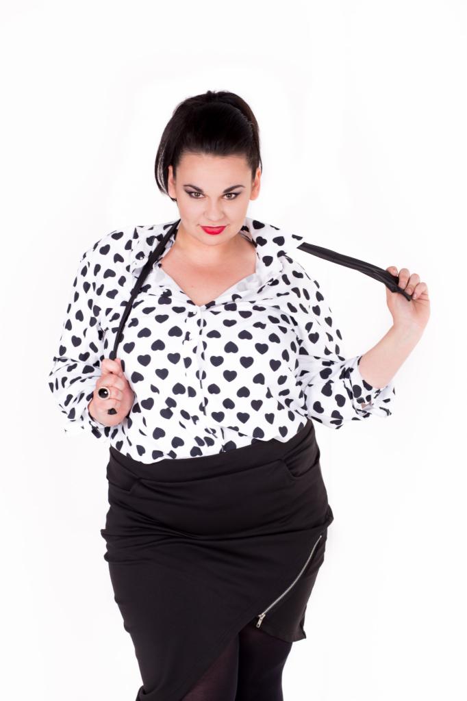 Kobieta w bluzce w czarne serca i czarnej spódnicy z asymetrycznym zamkiem, pozująca na białym tle.