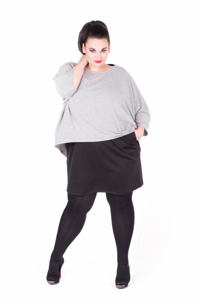 Kobieta w szarej bluzce typu nietoperz i czarnej spódnicy z kieszenią, pozuje na białym tle, prezentując stylizację plus size.