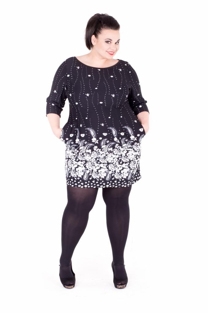 Kobieta w czarnej sukience z białym wzorem paisley, ciemne rajstopy i czarne buty na platformie, pozująca na białym tle.