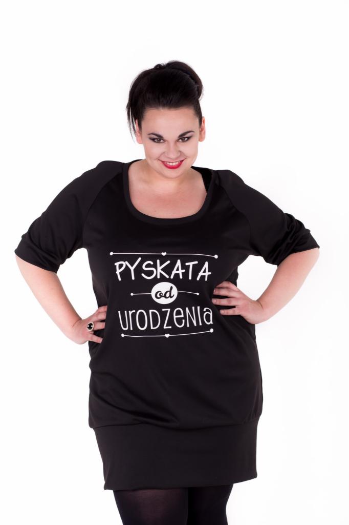 Kobieta pozuje w czarnej tunice z nadrukiem 'Pyskata od urodzenia', ręce oparte na biodrach, pierścionek na palcu.