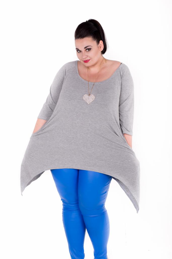 Kobieta w szarej tunice z kieszeniami i niebieskich, błyszczących legginsach na białym tle, z naszyjnikiem w kształcie serca.