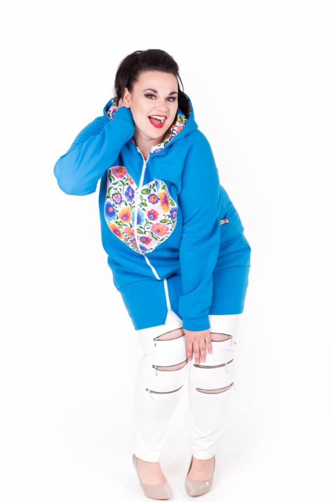 Uśmiechnięta kobieta w niebieskiej bluzie z kapturem, z folkowym wzorem w kształcie serca, białych spodniach z zamkami na kolanach i beżowych szpilkach, pozuje na białym tle.