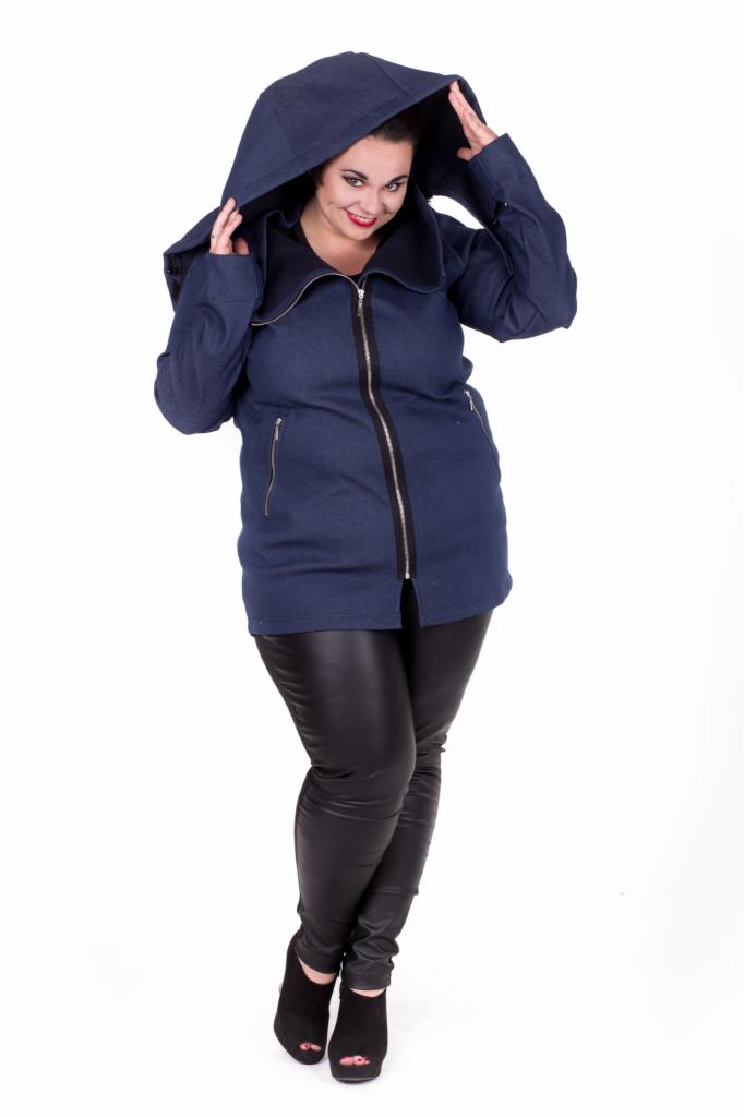 Kobieta w granatowej kurtce z kapturem, zapinanej na zamek błyskawiczny, pozuje na białym tle, ubrana w czarne, skórzane legginsy i czarne buty na platformie.