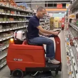 Operator na czerwonym, samojezdnym urządzeniu czyszczącym, poruszający się między regałami sklepowymi z artykułami spożywczymi.