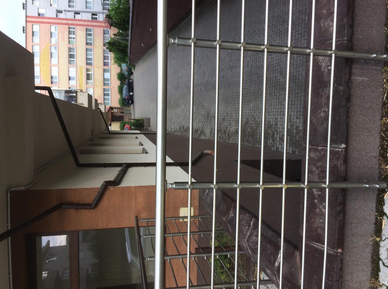 Nowoczesna, metalowa balustrada balkonowa na tle budynku wielorodzinnego i brukowanego chodnika.