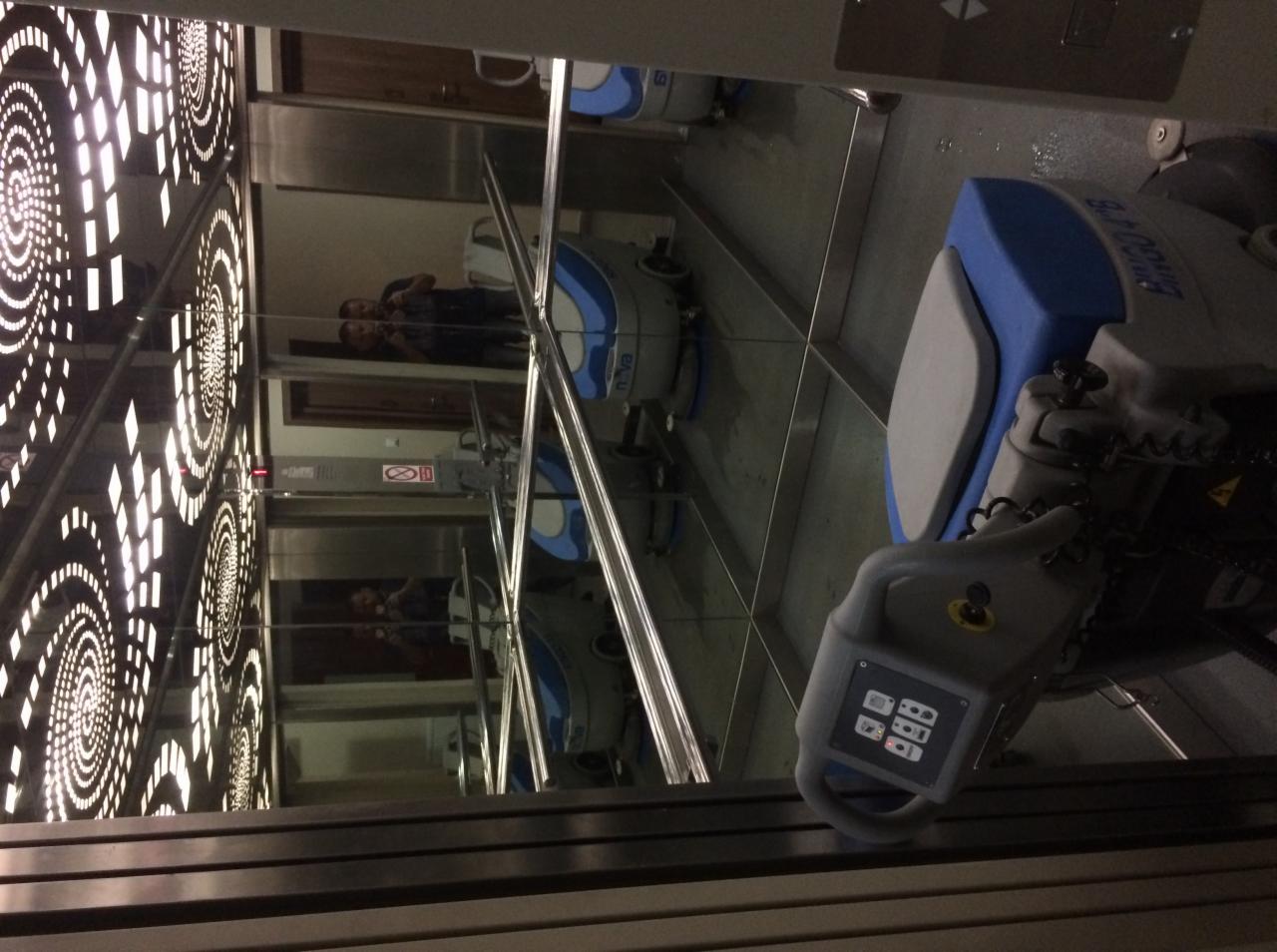 Automat do mycia podłóg w windzie z lustrzanymi ścianami, widoczne odbicie dwóch osób i wzorzysty sufit.