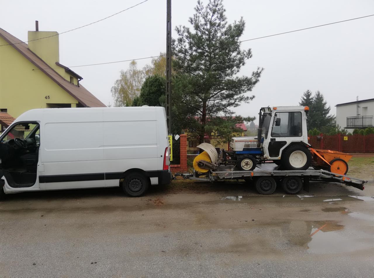Biały bus z otwartymi drzwiami i przyczepa z małym traktorem do odśnieżania na tle zabudowy jednorodzinnej w pochmurny dzień.