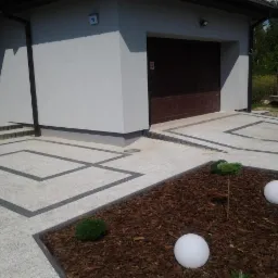 Jasnoszara kostka brukowa z ciemnymi obramowaniami tworzącymi geometryczne wzory na podjeździe, rabata z korą, kuliste lampy ogrodowe i małe krzewy, garaż w tle.