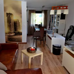 Jasne biuro z drewnianą podłogą, brązową sofą i plakatem z motywem miejskim. Elementy wystroju: białe biurko, krzesła, lampa i stolik z dekoracją.