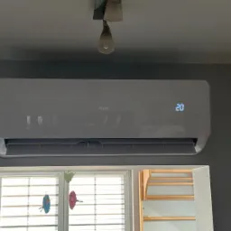 Biały klimatyzator marki AUX zamontowany na szarej ścianie, ustawiony na 20 stopni, widoczne okno z dekoracjami i fragment schodów w tle.