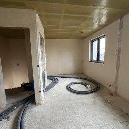 Comfort System House Tomasz Pawłowski - Wnętrze w stanie surowym w Ostródzie z widocznymi przewodami instalacji grzewczej na podłodze i izolacją na suficie, przygotowane do dalszych prac wykończeniowych.