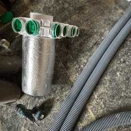 Comfort System House Tomasz Pawłowski - Elementy instalacji rekuperacji z rozdzielaczem, rurami karbowanymi i wiertarką na surowym podłożu w trakcie montażu.