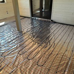Comfort System House Tomasz Pawłowski - Rozłożona instalacja ogrzewania podłogowego na folii z czerwoną kratką w jasnym pomieszczeniu z drewnianą boazerią i słupem konstrukcyjnym.
