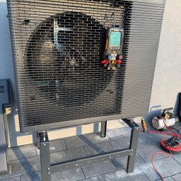 Comfort System House Tomasz Pawłowski - Czarna pompa ciepła Panasonic Aquarea na metalowym stelażu, podłączona do miernika ciśnienia i pompy próżniowej podczas instalacji na zewnątrz budynku.