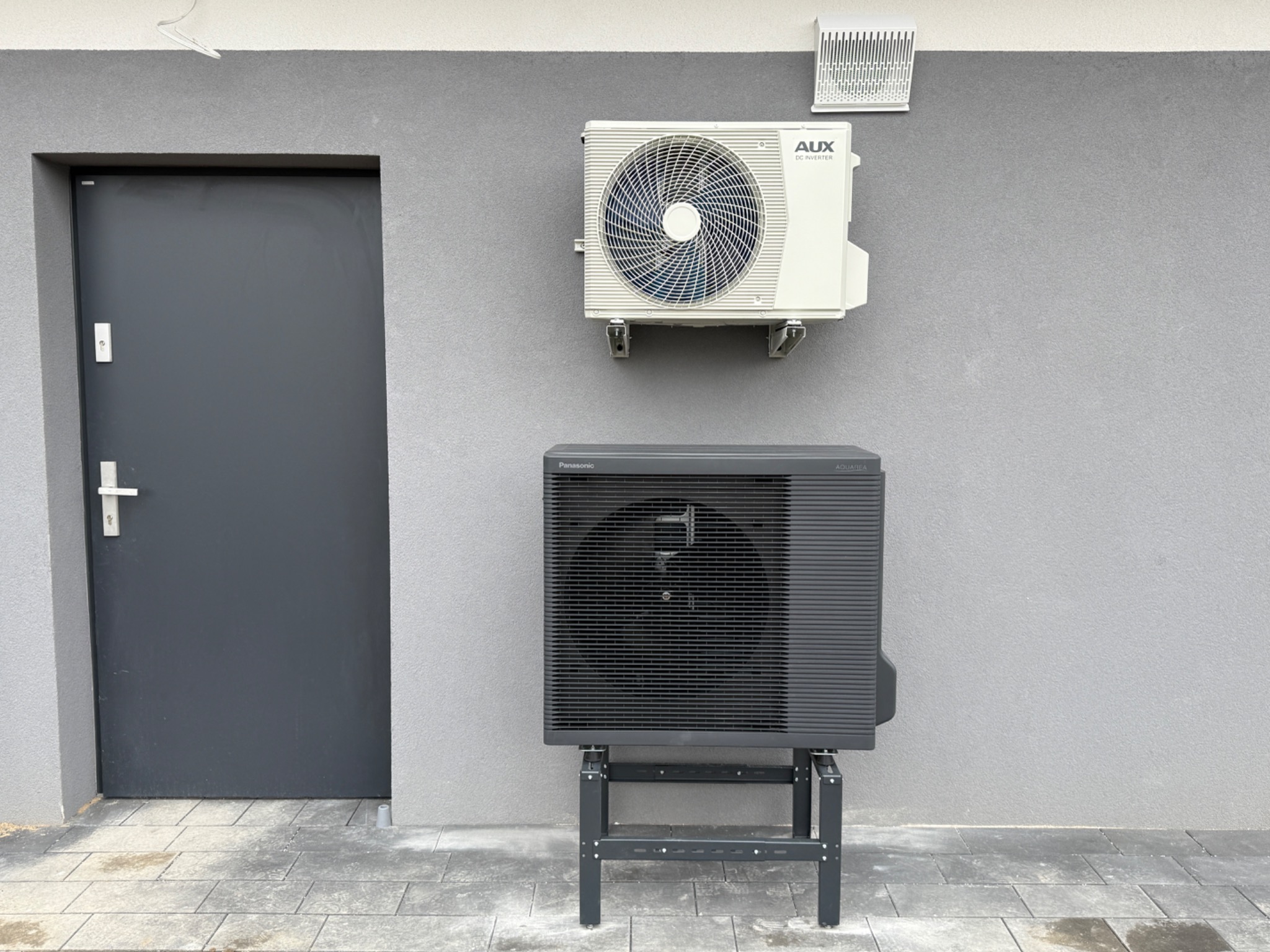 Dwie jednostki klimatyzacyjne zamontowane na szarej ścianie zewnętrznej budynku obok ciemnoszarych drzwi wejściowych; górna jednostka marki AUX, dolna - Panasonic Aquarea, obie z widocznymi...