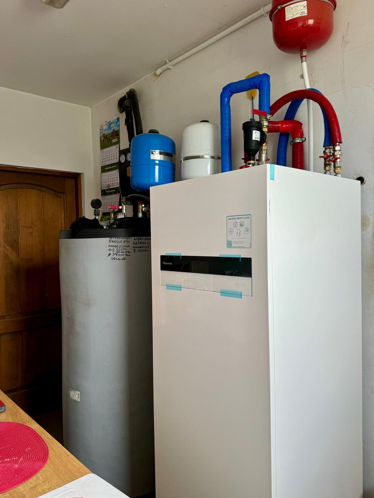 Biała pompa ciepła Panasonic Aquarea Smart Cloud z niebieskimi i czerwonymi rurami, zbiornik szary z notatkami, kalendarz na ścianie, w pomieszczeniu technicznym.