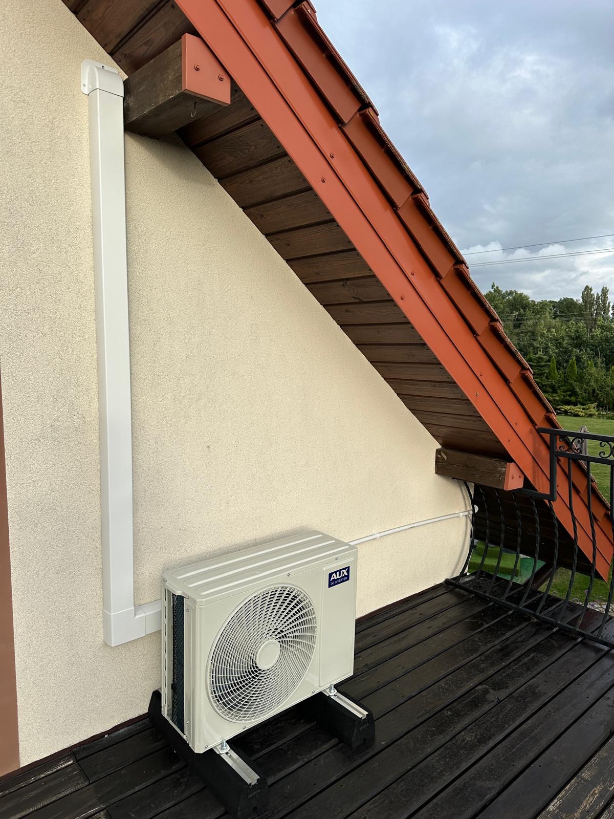 Zewnętrzna jednostka klimatyzacji marki AUX zamontowana na balkonie z drewnianą podłogą, z widoczną rurą odprowadzającą skropliny i fragmentem dachu z brązową dachówką.