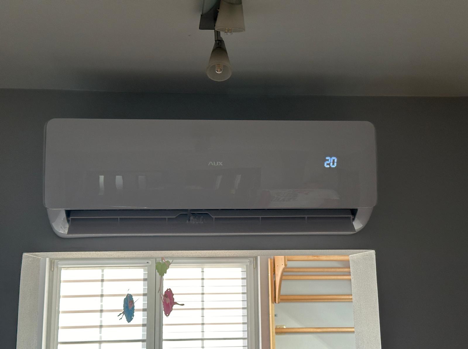Biały klimatyzator marki AUX zamontowany na szarej ścianie, ustawiony na 20 stopni, widoczne okno z dekoracjami i fragment schodów w tle.