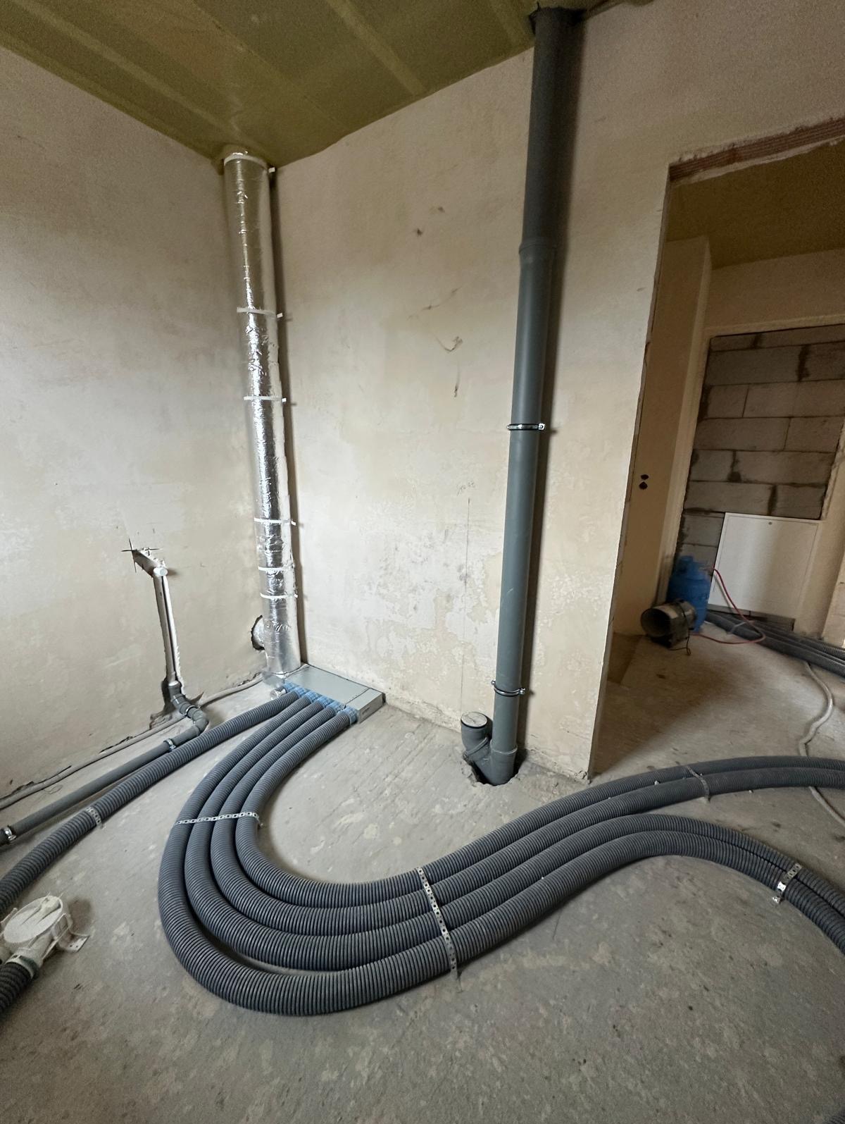 Szare karbowane rury wentylacyjne ułożone na surowym betonowym podłożu, prowadzące do pionowych rur wentylacyjnych, jedna izolowana folią aluminiową, w surowym wnętrzu budynku w trakcie instalacji.