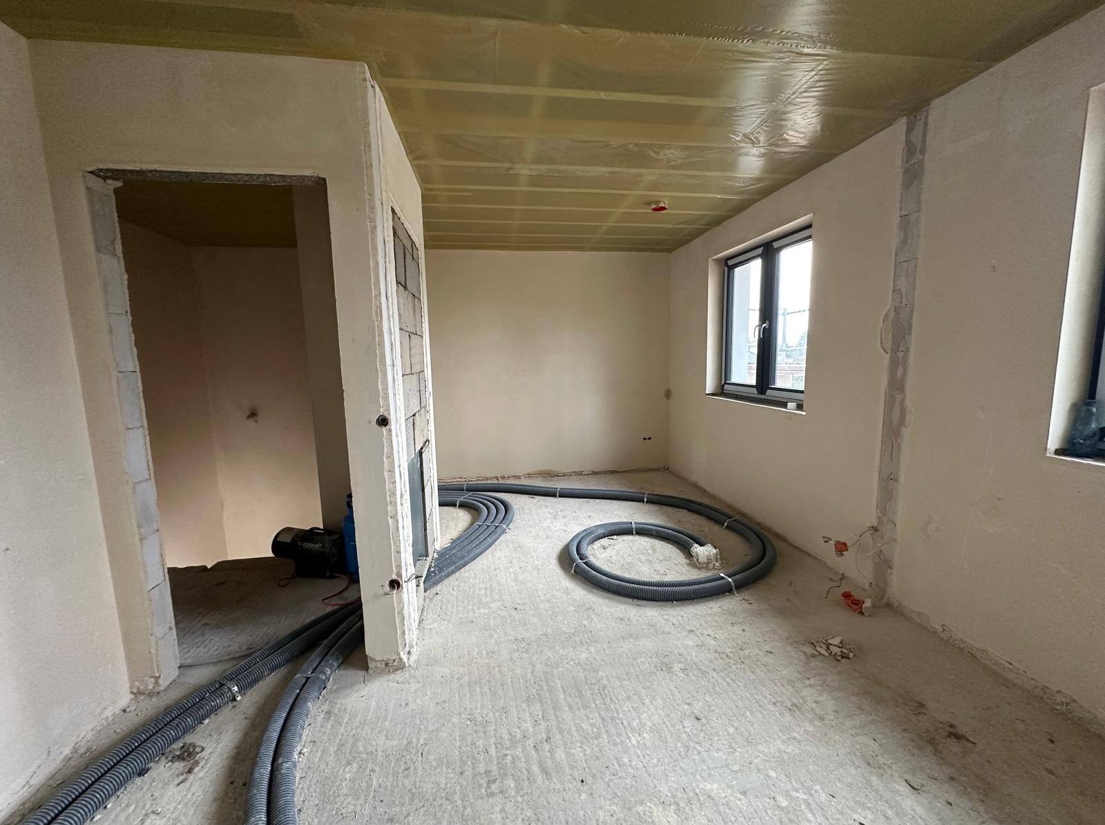 Wnętrze w stanie surowym w Ostródzie z widocznymi przewodami instalacji grzewczej na podłodze i izolacją na suficie, przygotowane do dalszych prac wykończeniowych.