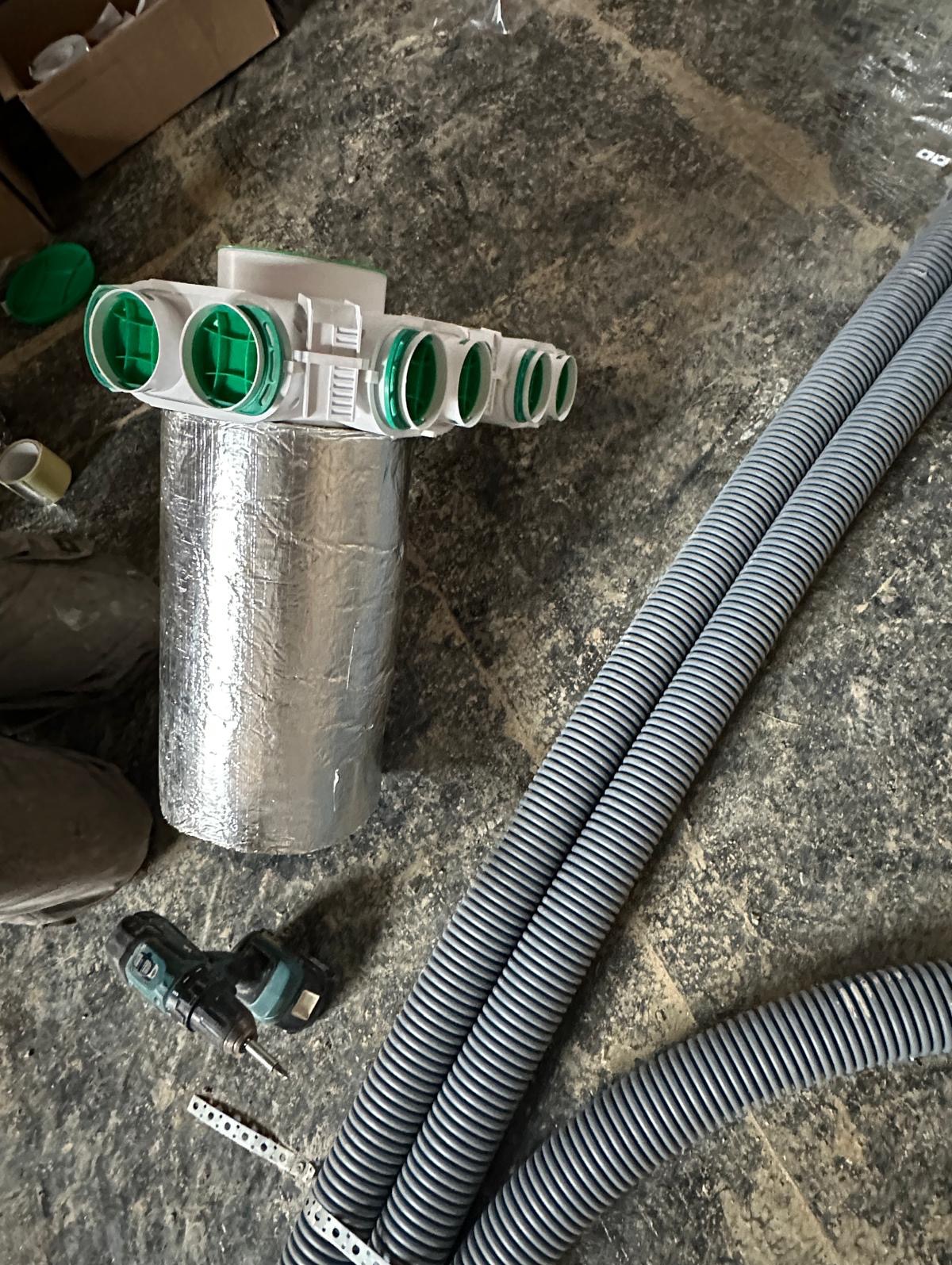 Elementy instalacji rekuperacji z rozdzielaczem, rurami karbowanymi i wiertarką na surowym podłożu w trakcie montażu.