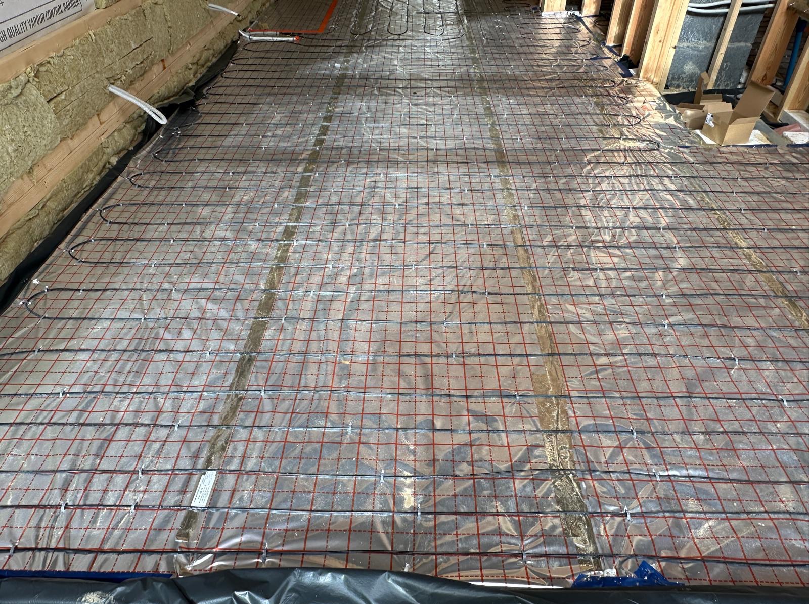 Rozłożona mata grzewcza na folii z siatką, przygotowana do zalania wylewką, widoczne kable zasilające, ocieplenie stropu wełną mineralną.