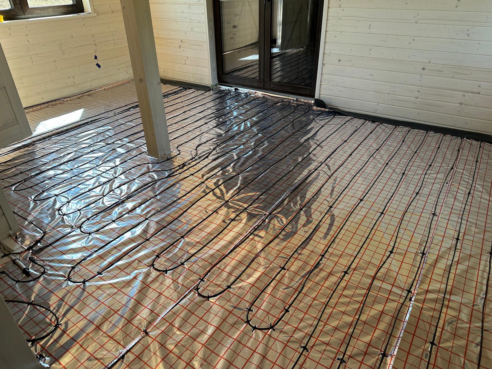 Rozłożona instalacja ogrzewania podłogowego na folii z czerwoną kratką w jasnym pomieszczeniu z drewnianą boazerią i słupem konstrukcyjnym.