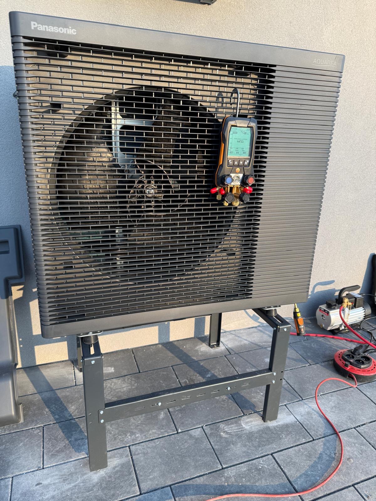 Czarna pompa ciepła Panasonic Aquarea na metalowym stelażu, podłączona do miernika ciśnienia i pompy próżniowej podczas instalacji na zewnątrz budynku.