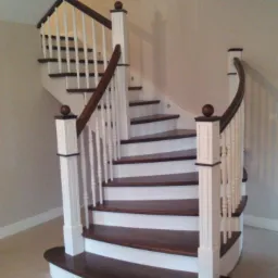 Kręte schody drewniane z białymi balustradami i ciemnobrązowymi stopniami, zakończone ozdobnymi kulami na słupkach, w jasnym wnętrzu.