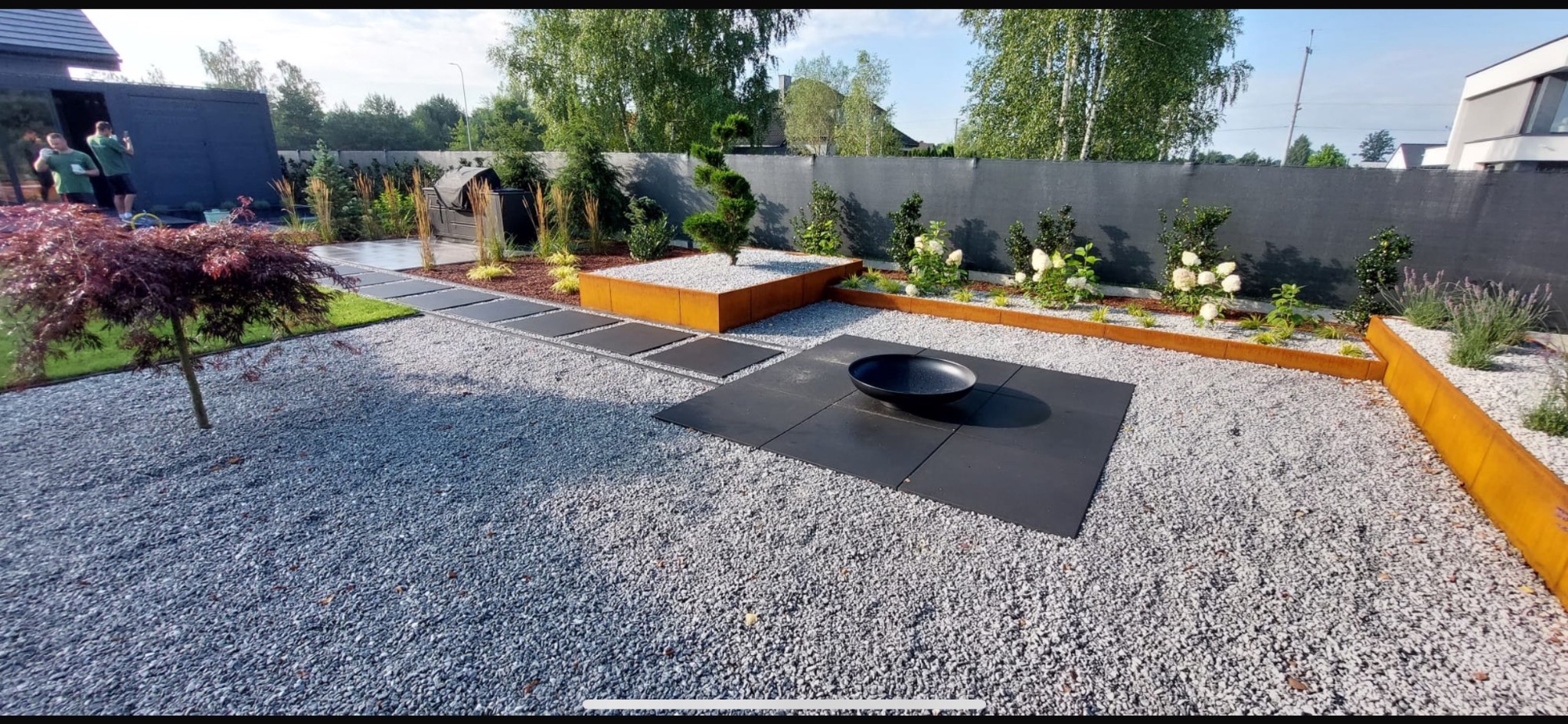 Nowoczesny ogród z paleniskiem, ścieżką z płyt i rabatami z roślinami w metalowych obrzeżach. Szara nawierzchnia żwirowa, minimalistyczny design, widok z perspektywy szerokokątnej.