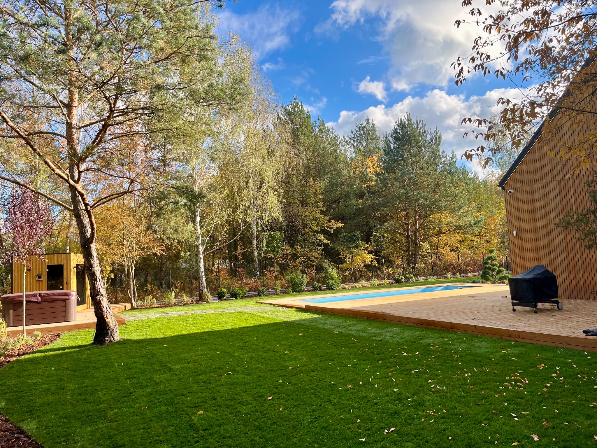 Nowoczesny ogród z basenem i drewnianym tarasem w jesiennej scenerii. Widoczna sauna i grill. Starannie utrzymany trawnik z opadłymi liśćmi. Błękitne niebo z chmurami w tle.