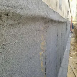 Szara izolacja pionowa fundamentu budynku z widoczną fakturą, zabezpieczona przed wilgocią, z wykopem wzdłuż ściany. Fragment budynku w tle.