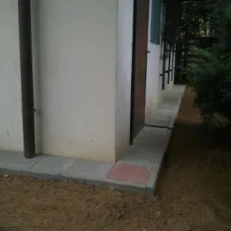 Ułożona kostka brukowa wzdłuż ściany budynku, z widoczną rurą spustową i fragmentem zieleni. Kostka szara, z pojedynczym elementem w kolorze ceglastym.